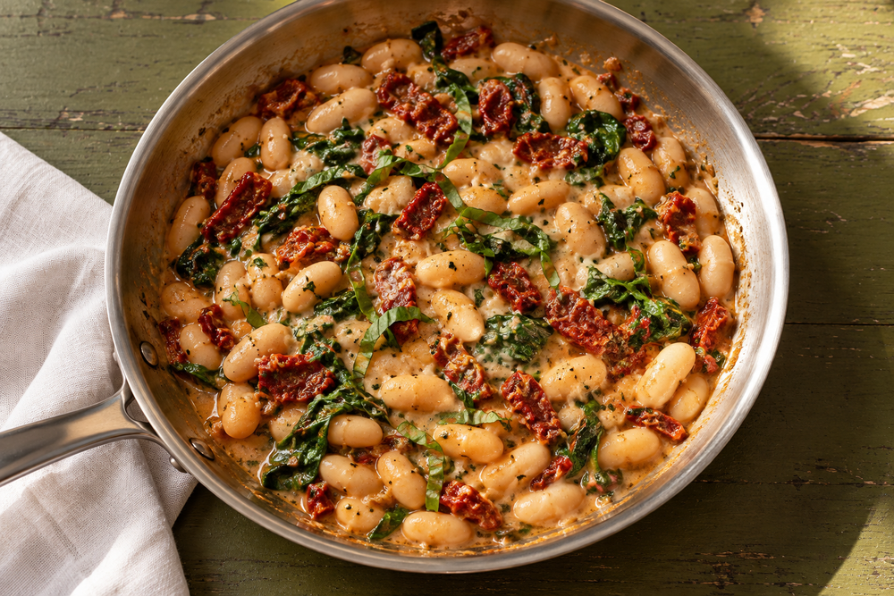 Cannellini beans with sun-dried tomatoes, spinach and basil in a stainless steel skillet on an olive table.