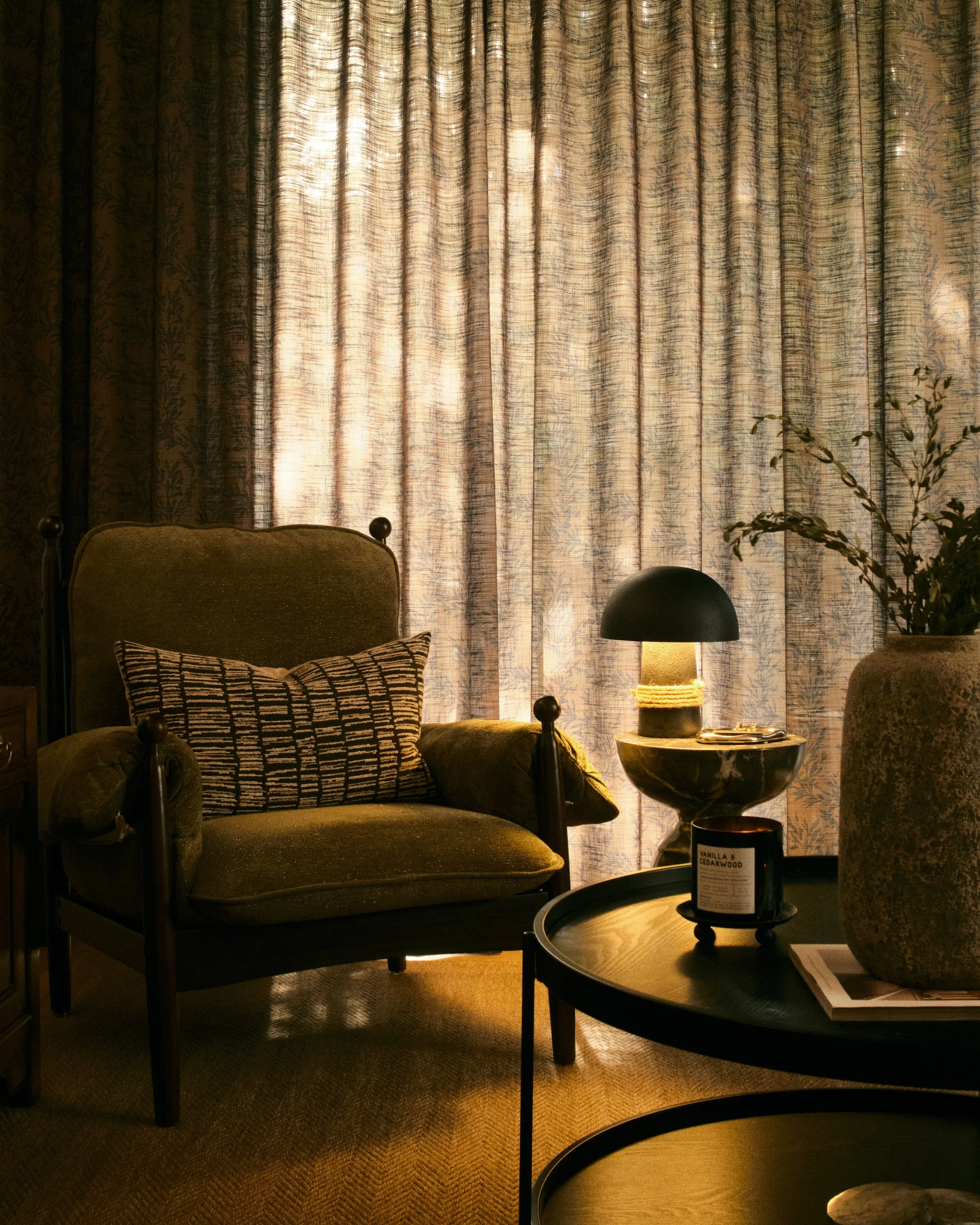 Olive green velvet armchair with textured throw pillow beside a marble side table with mushroom lamp and cedarwood candle, warm ambient interior lighting