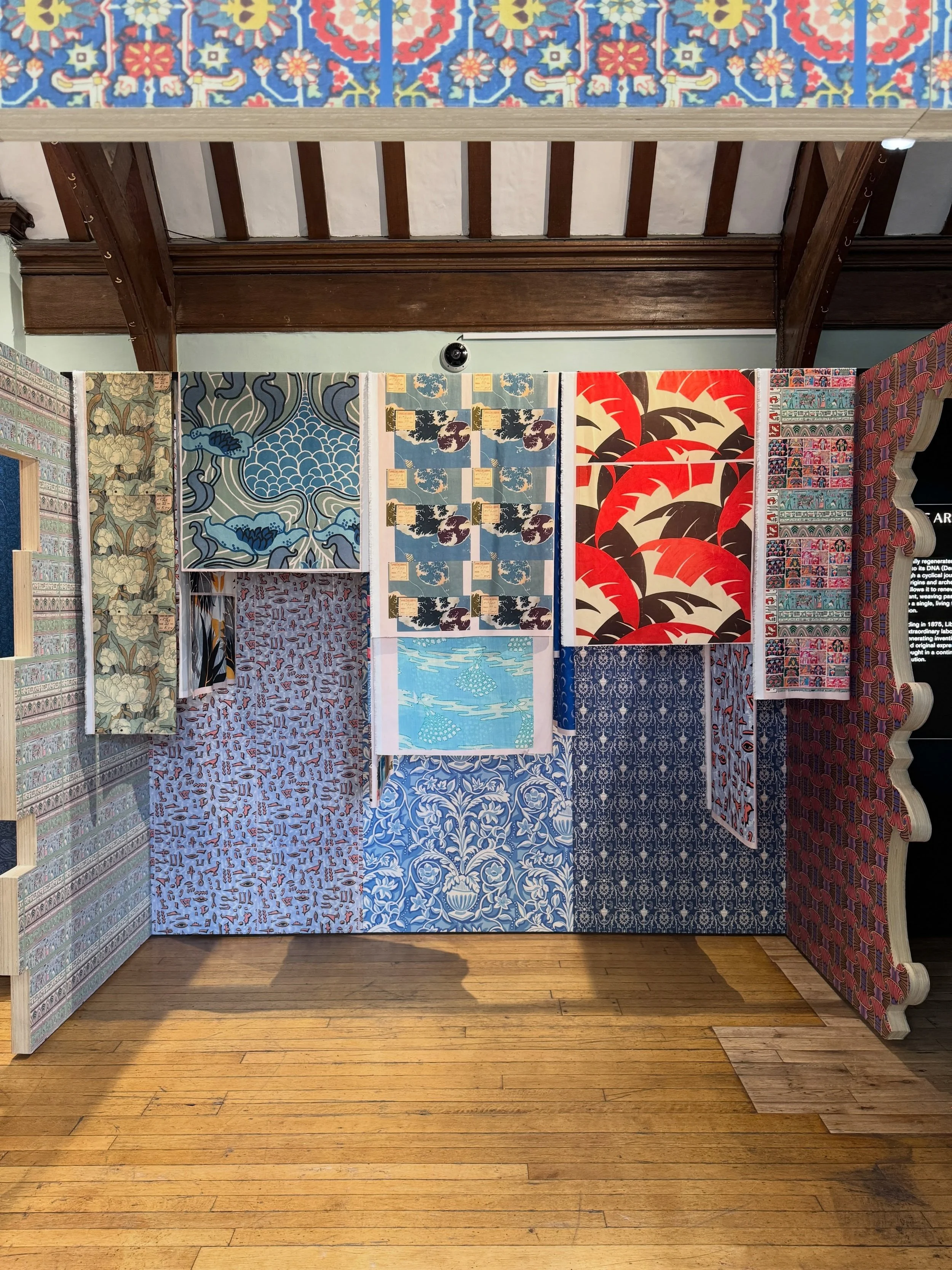 Printed textiles hanging against patterned blue wallpaper and timber beams at Liberty London.
