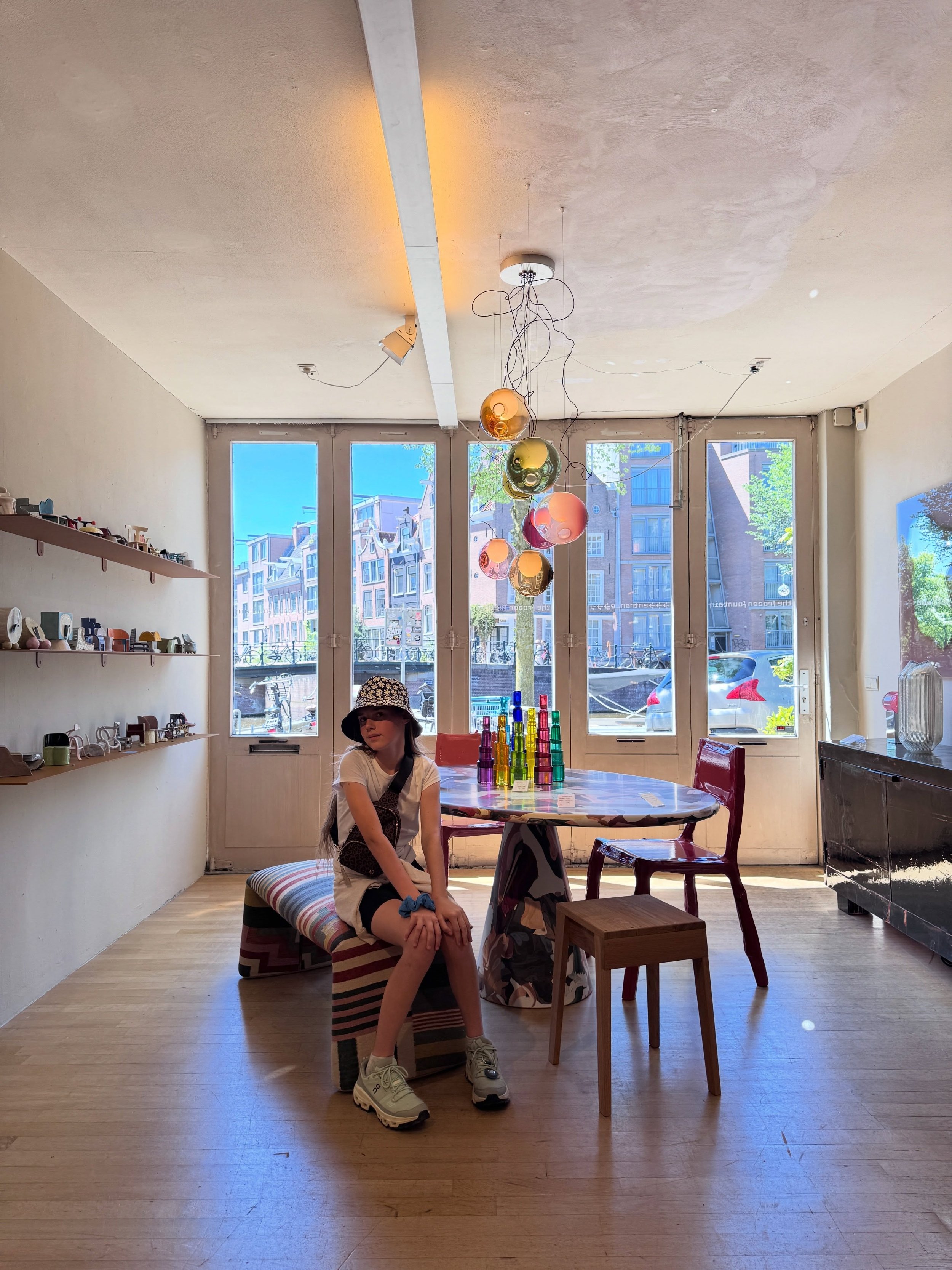 Amsterdam design showroom with a sculptural pendant, wood table, chairs, wall shelving, and a seated visitor.