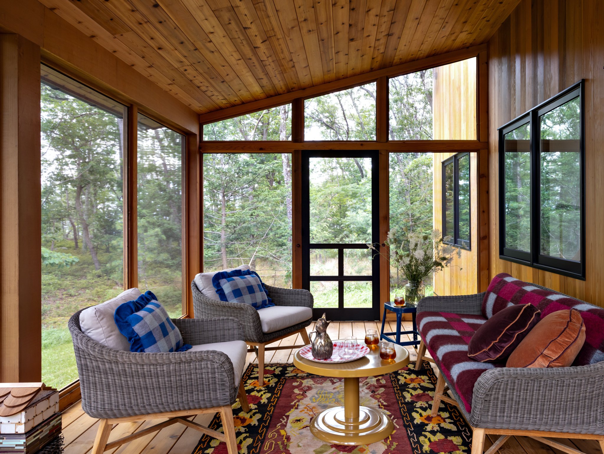 Screened porch with a timber ceiling, patterned seating, blue checked cushions, a yellow side table, and woodland views.