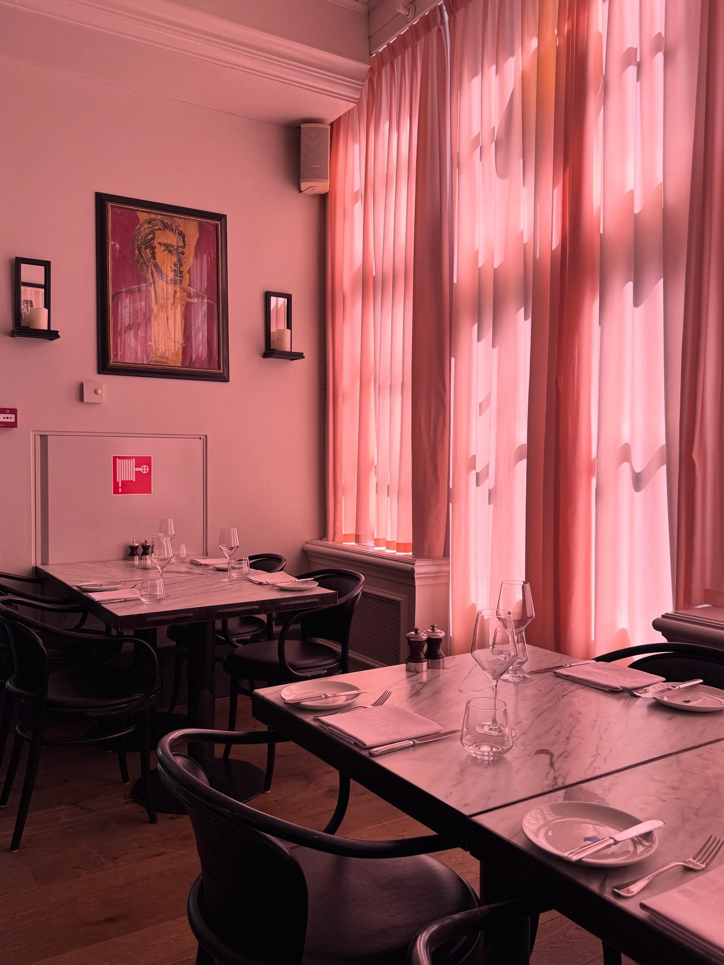 Pink dining room in Amsterdam with sheer curtains, framed artwork, dark tables, and upholstered seating.