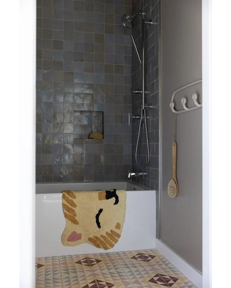 Glazed Green Tile Shower | Amy Pigliacampo Interiors Bathtub and shower with glazed green wall tile, dark fixtures, a patterned towel, and multicolored geometric floor tile.
