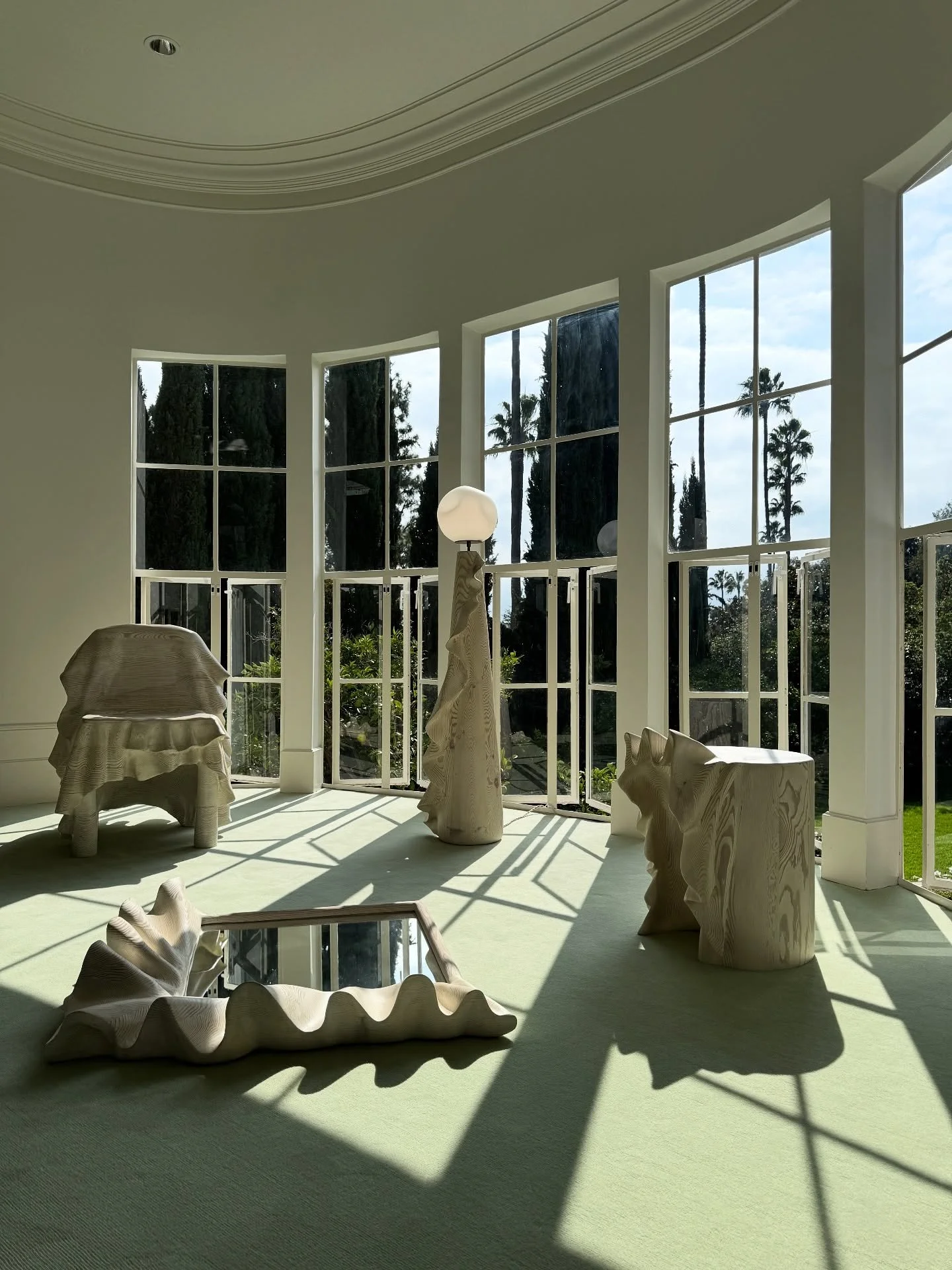 Sunlit gallery room with tall black-framed windows, curved white sofas, and sculptural furnishings.