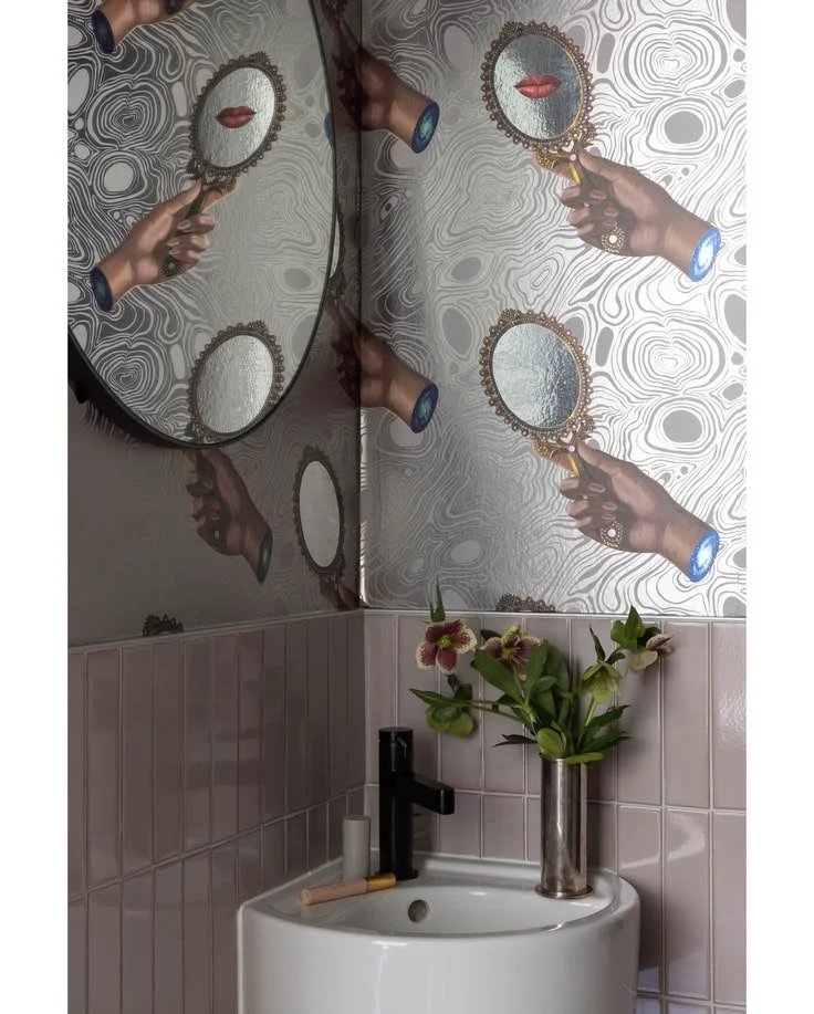 Powder room with illustrated hand-and-mirror wallpaper, pale pink tile, a white corner sink, and dark wall faucet.