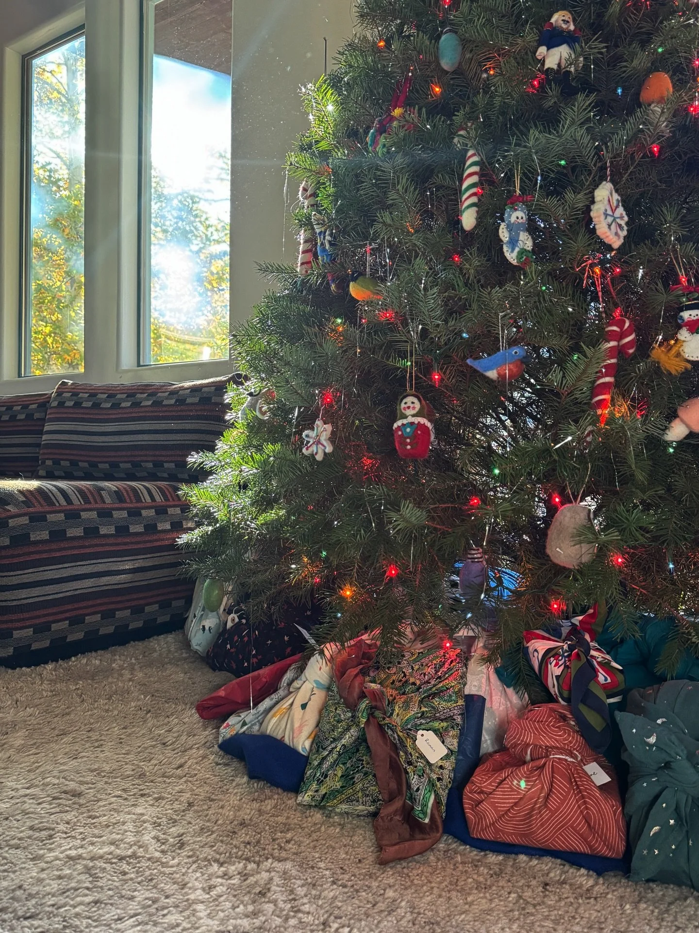 Christmas tree beside a window with fabric-wrapped gifts and patterned upholstery nearby.
