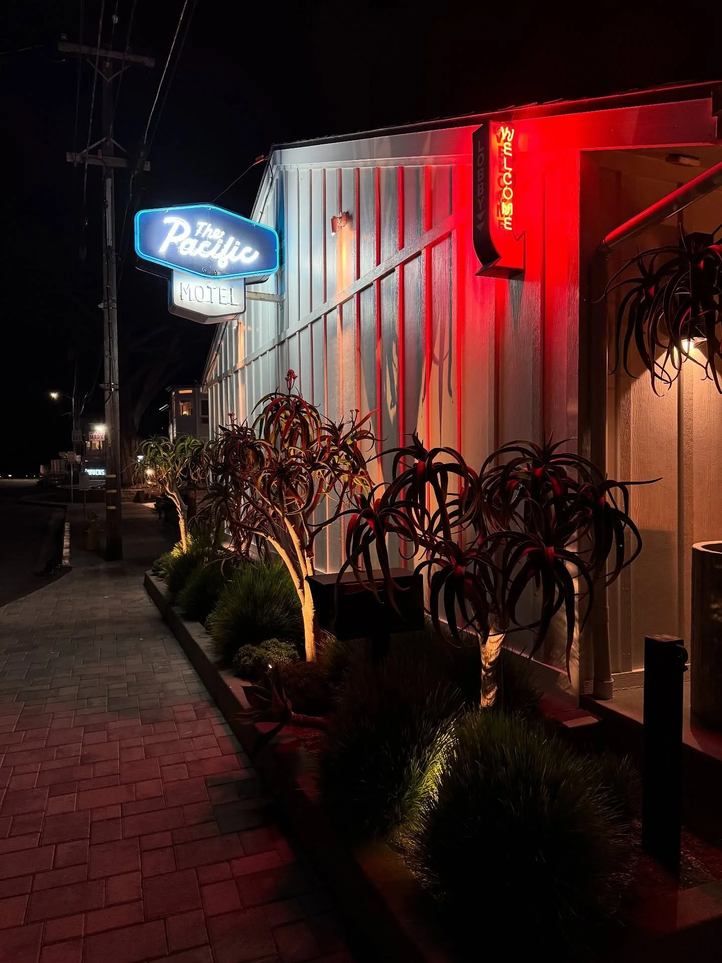 The Pacific Motel exterior at night, with illuminated signs, warm red lighting, and planted borders.