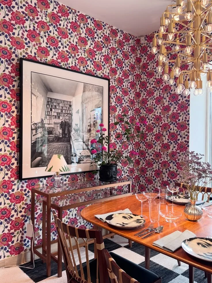 Dining table set with orange placemats beneath a brass chandelier and red floral wallpaper at Svenskt Tenn.