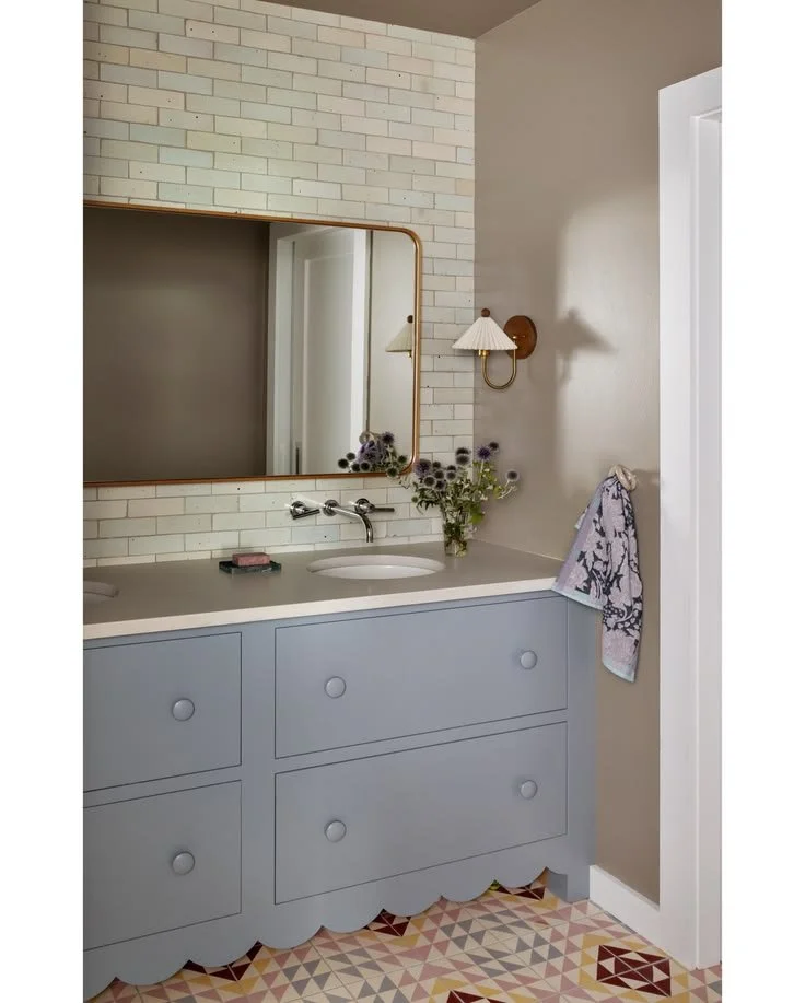Scalloped Vanity & Pale Green Tile | Amy Pigliacampo Interiors Bathroom with a blue-gray scalloped vanity, chrome wall faucets, pale green tile, brass-framed mirror, and geometric floor tile.