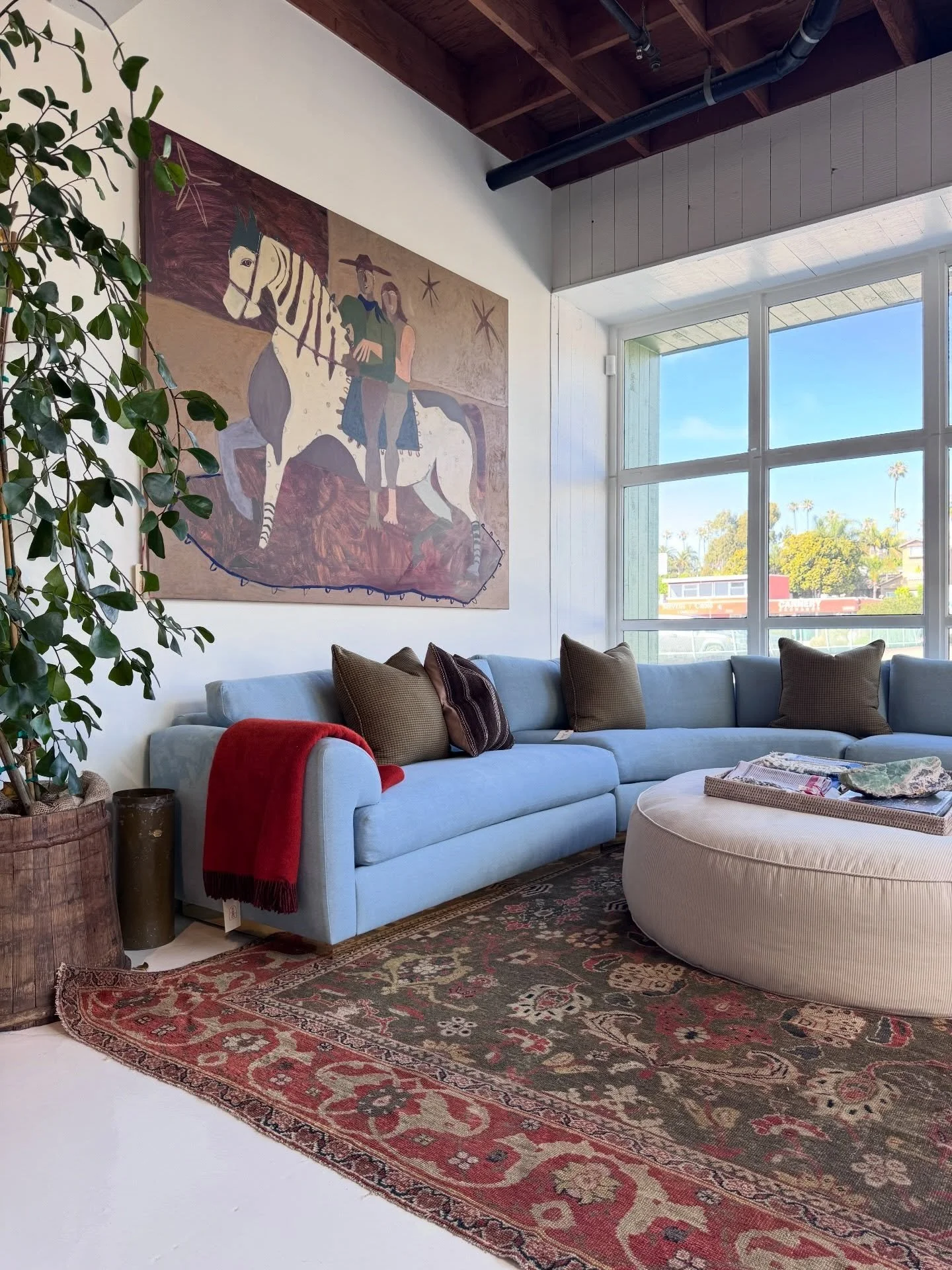 Shop interior with a pale sectional sofa, round ottoman, vintage rug, wall art, plants, and timber ceiling.