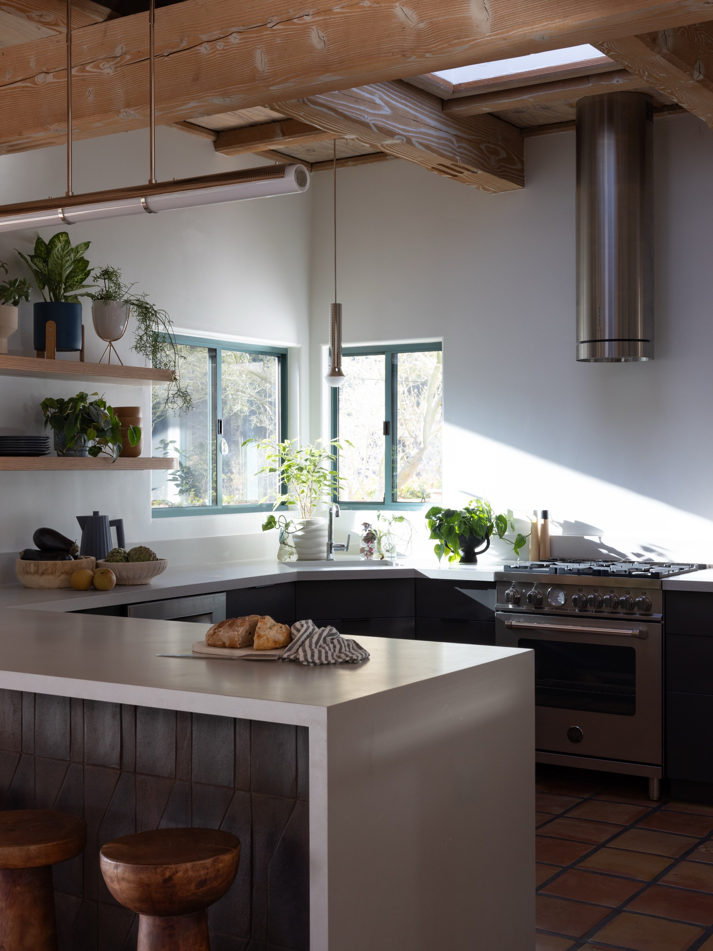 Kitchen with exposed wood beams, gray cabinetry, pale counters, open shelves, plants, and a sculptural stool.