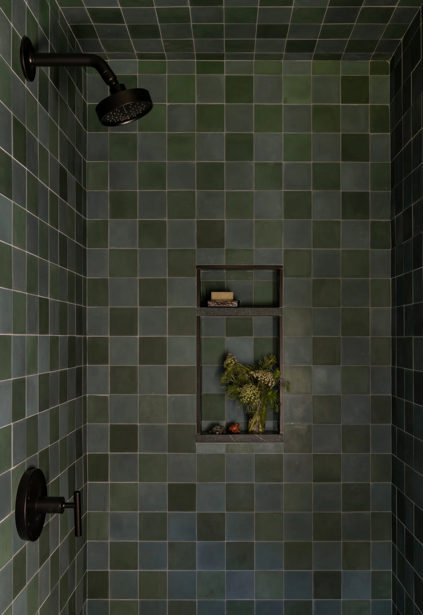Guest-house shower with mottled green square tile, dark fixtures, and a recessed niche lined with floral-patterned tile.