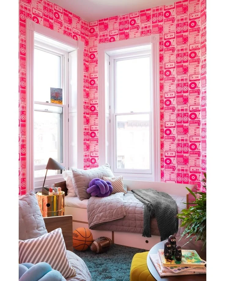 Bedroom with pink illustrated wallpaper, a corner window, layered pink and green bedding, and a multicolored rug.