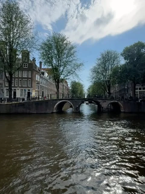 Amsterdam canal with a stone bridge, narrow houses, and trees along the water.