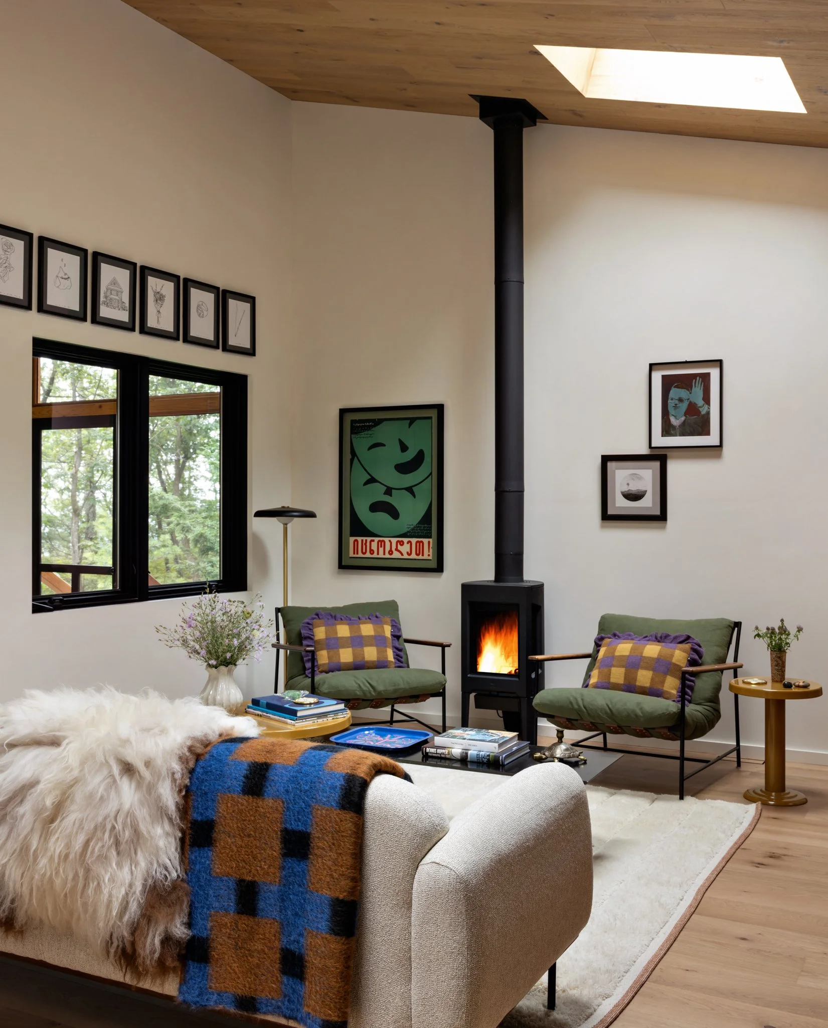 Guest-house living room with a black wood stove, cream sofa, blue-and-ochre throw, patterned chairs, and a timber ceiling.