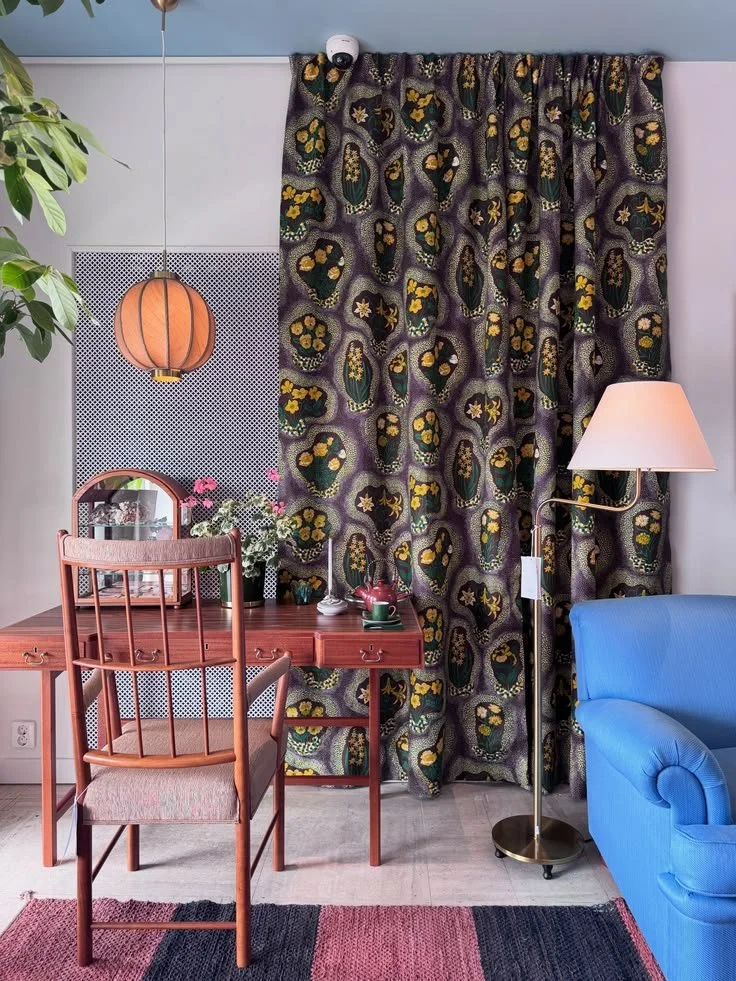Dining table with red chairs, dark botanical curtains, striped rug, and blue sofa at Svenskt Tenn.