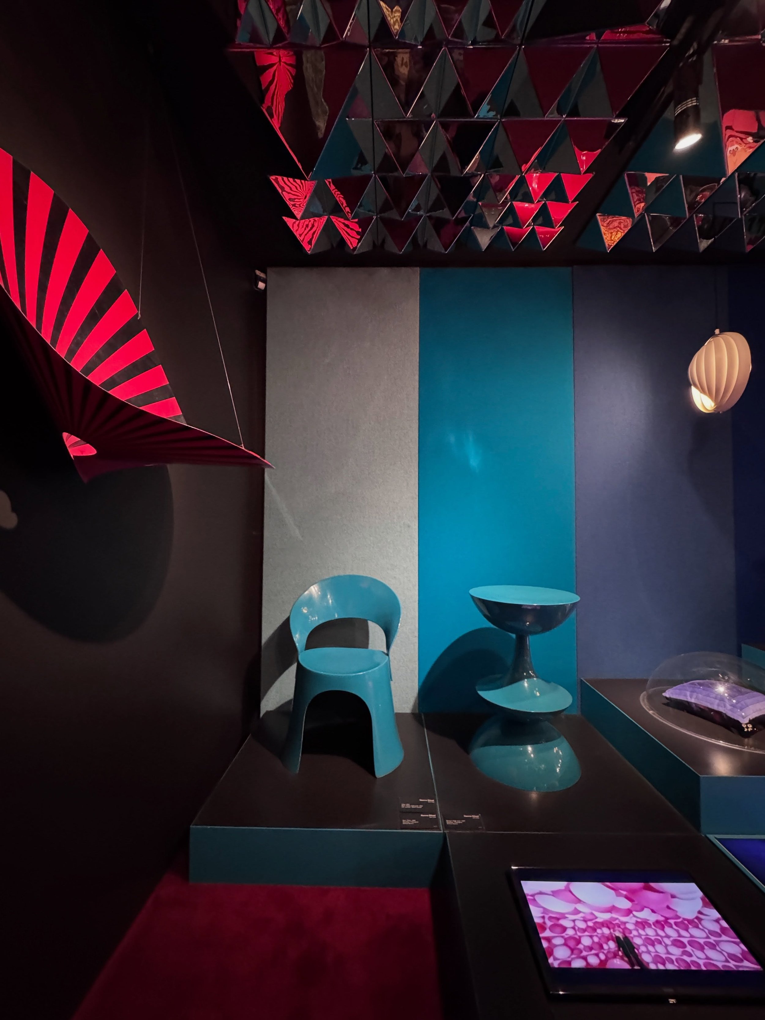 Two turquoise Panton chairs beneath a geometric ceiling installation, beside a red curtain.