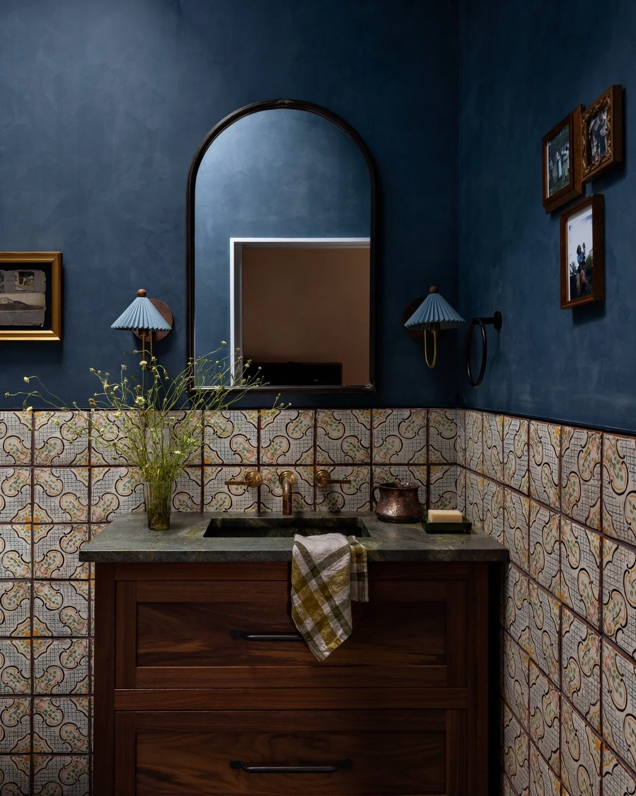 Bathroom with deep blue walls, an arched mirror, patterned tile, pleated sconces, and a wood vanity.