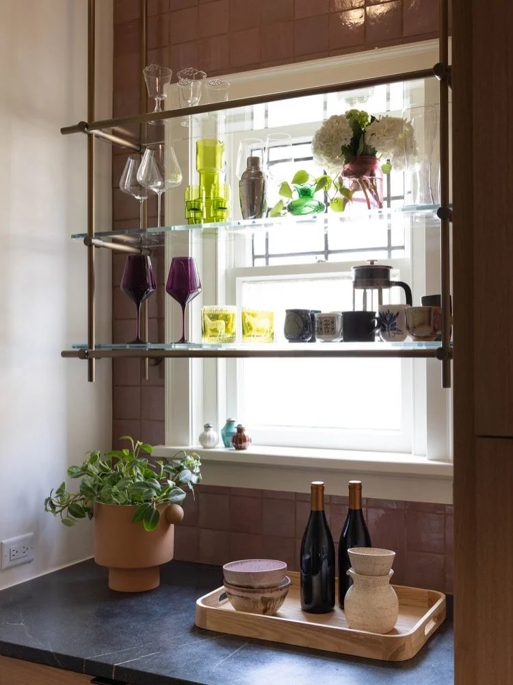 Brass-framed glass shelving across a kitchen window, with ceramics, plants, and a dark counter.