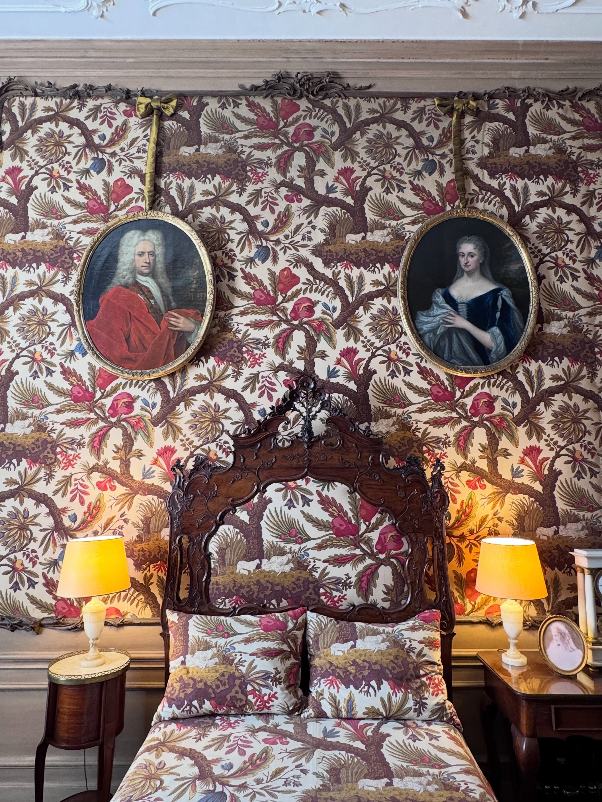 Historic Amsterdam bedroom with floral wallpaper, an ornate wood bed, and yellow-shaded bedside lamps.