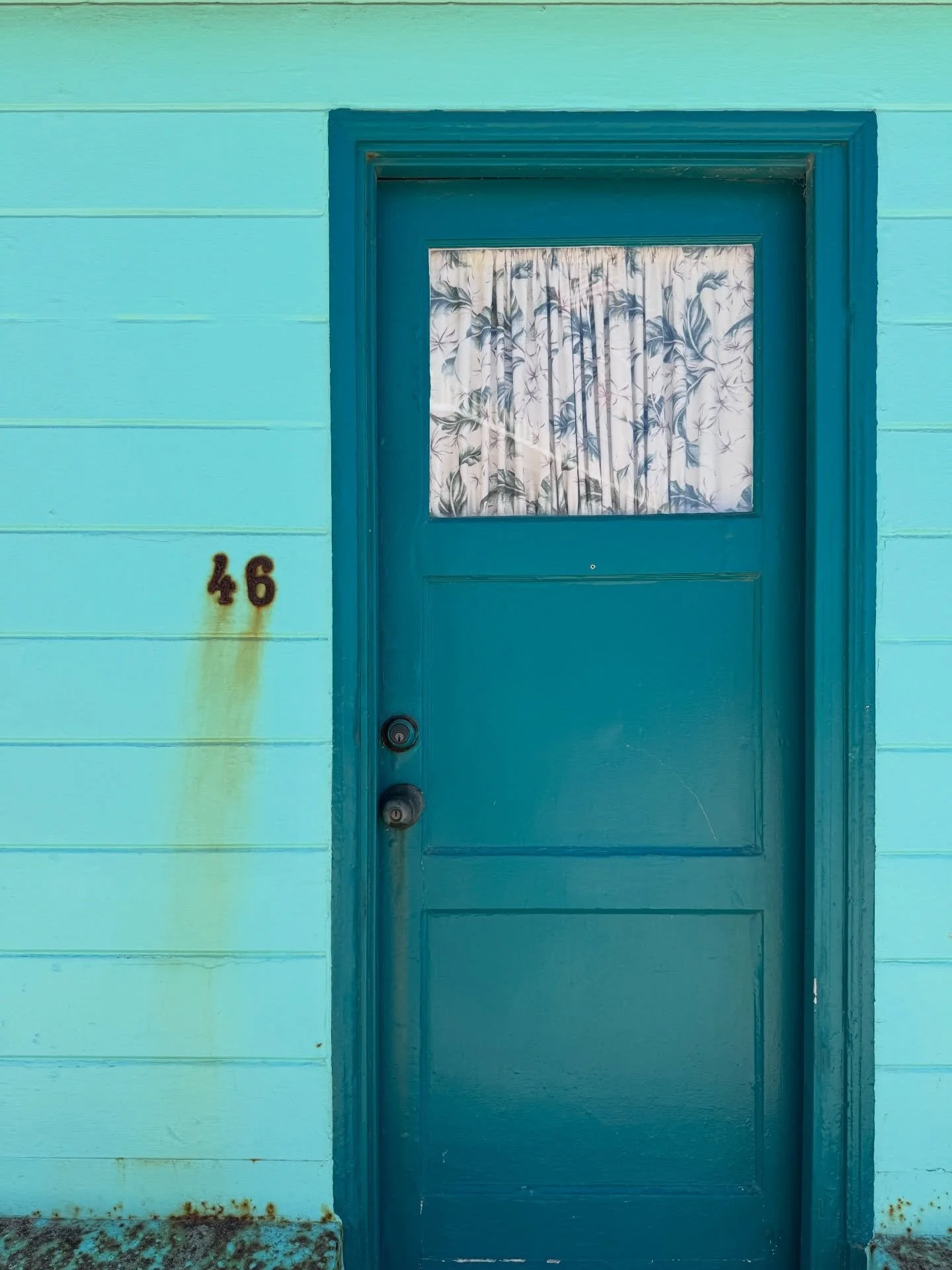 Turquoise cottage door with a small floral curtain and painted wood siding.
