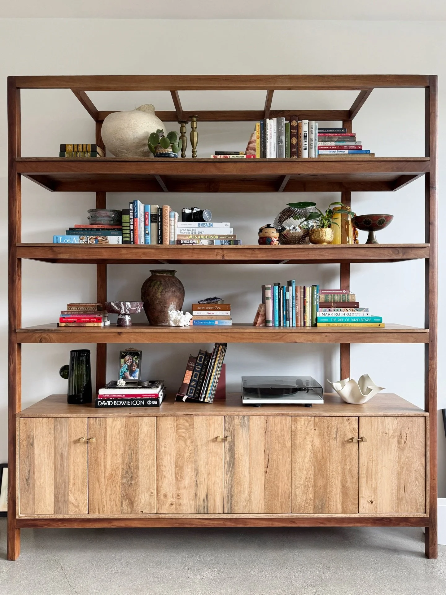 Wood display shelving above a low cabinet, with books, ceramics, framed art, and sculptural objects.