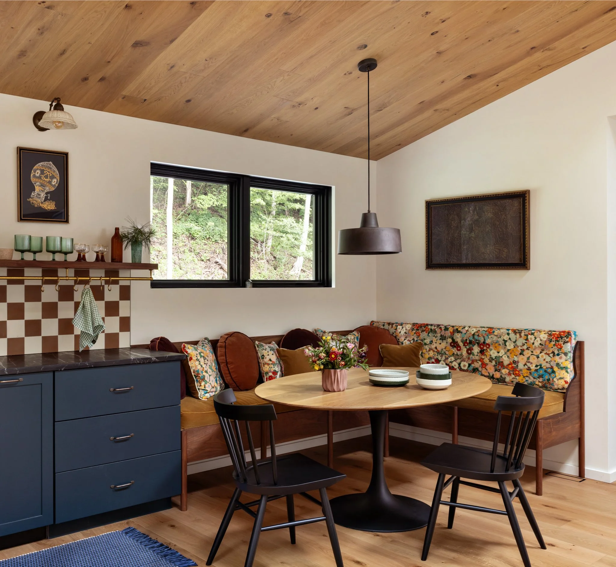 Guest-house dining nook with a round wood table, spindle chairs, patterned banquette cushions, and checkerboard kitchen tile.