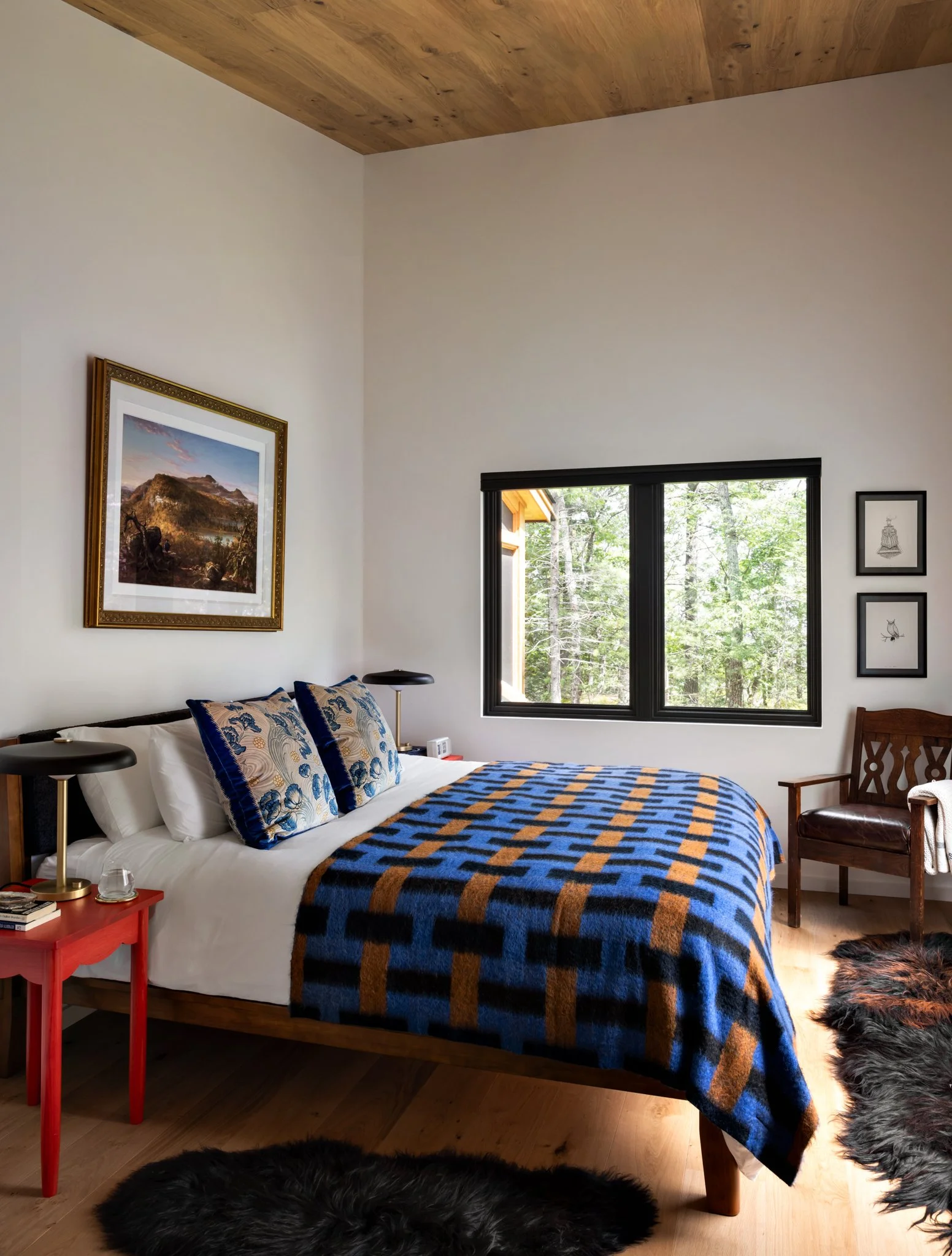 Guest-house bedroom with blue-and-ochre patterned bedding, a red bedside table, framed art, and black-trimmed windows.