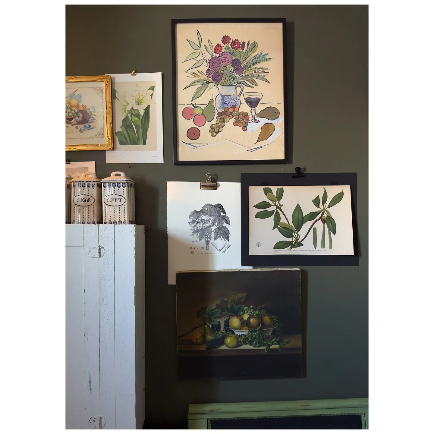 Botanical prints and framed paintings arranged on a dark wall beside a pale blue-gray cabinet.