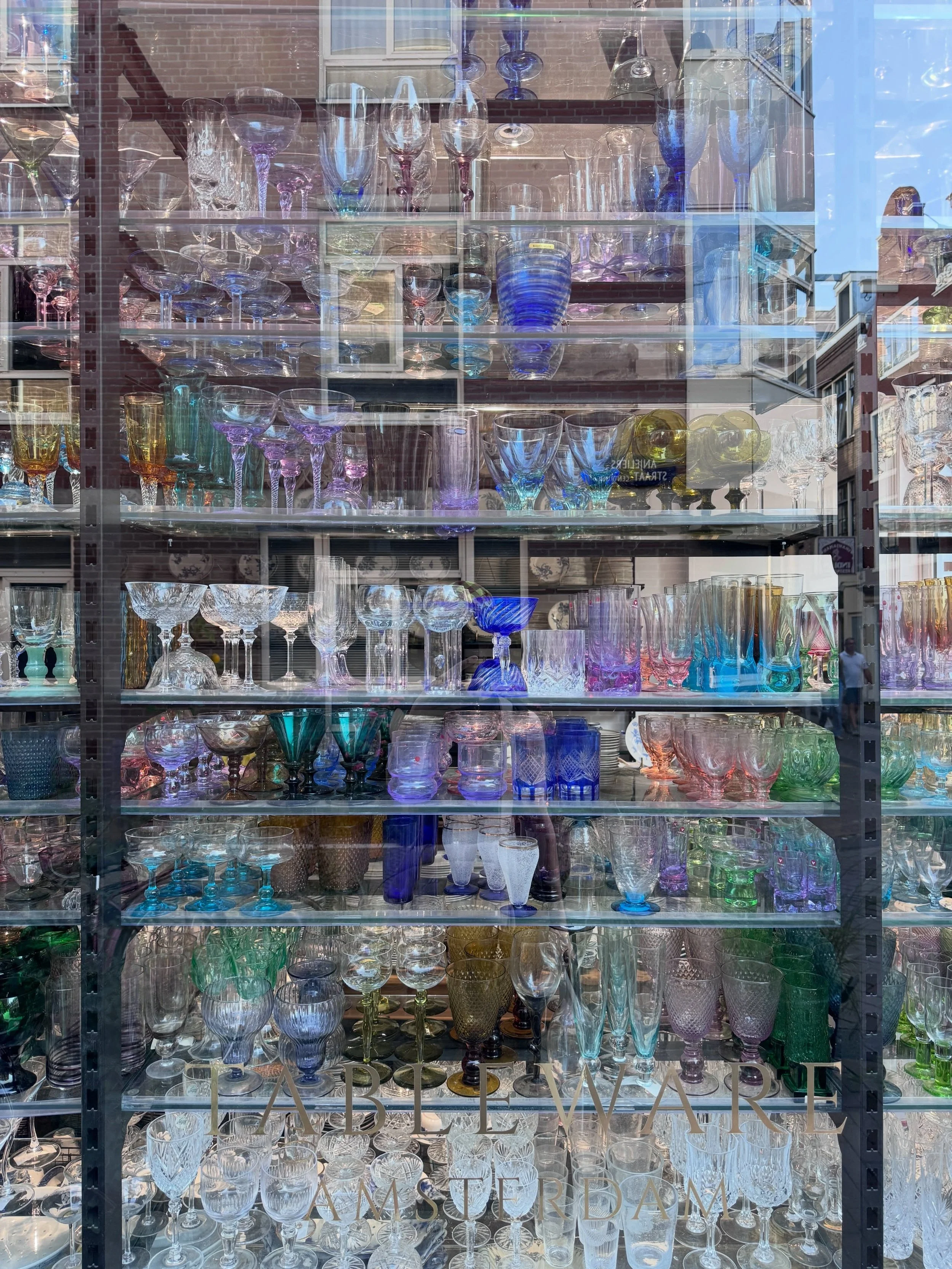 Colored glassware displayed in an Amsterdam shop window, with canal houses reflected in the glass.