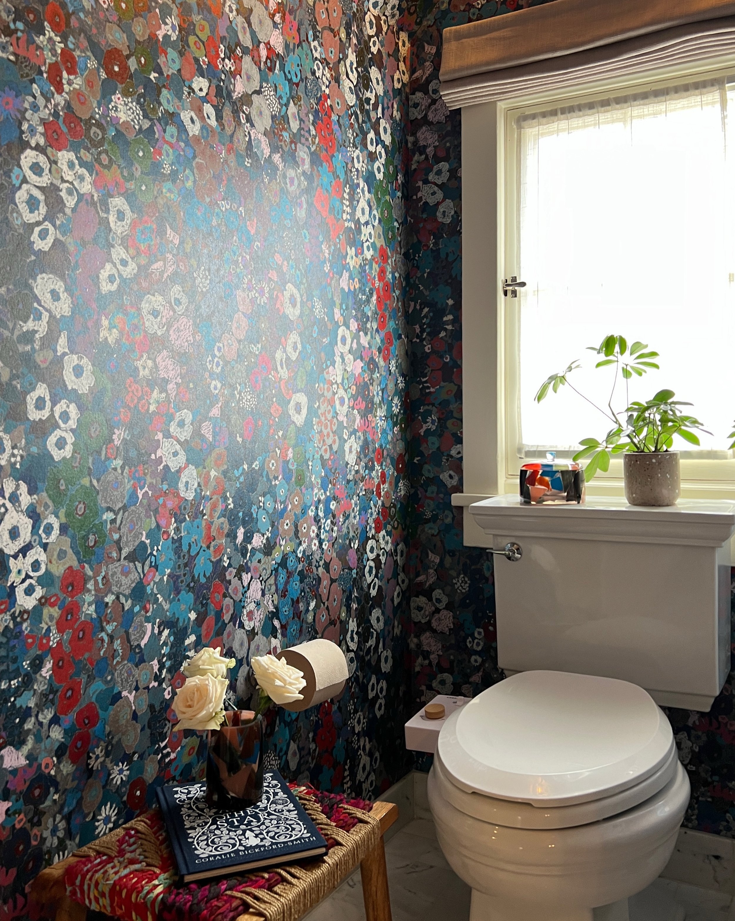 Powder room with multicolored floral wallpaper, a marble-topped wood cabinet, white toilet, and window.