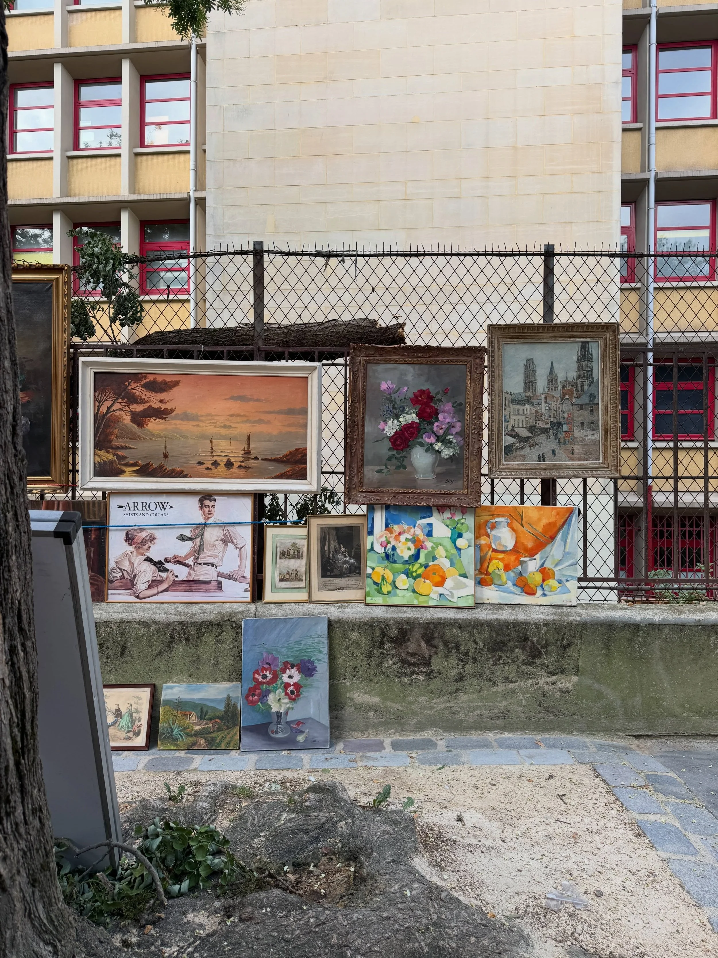 Framed paintings and small artworks arranged on outdoor shelves at a Paris market.