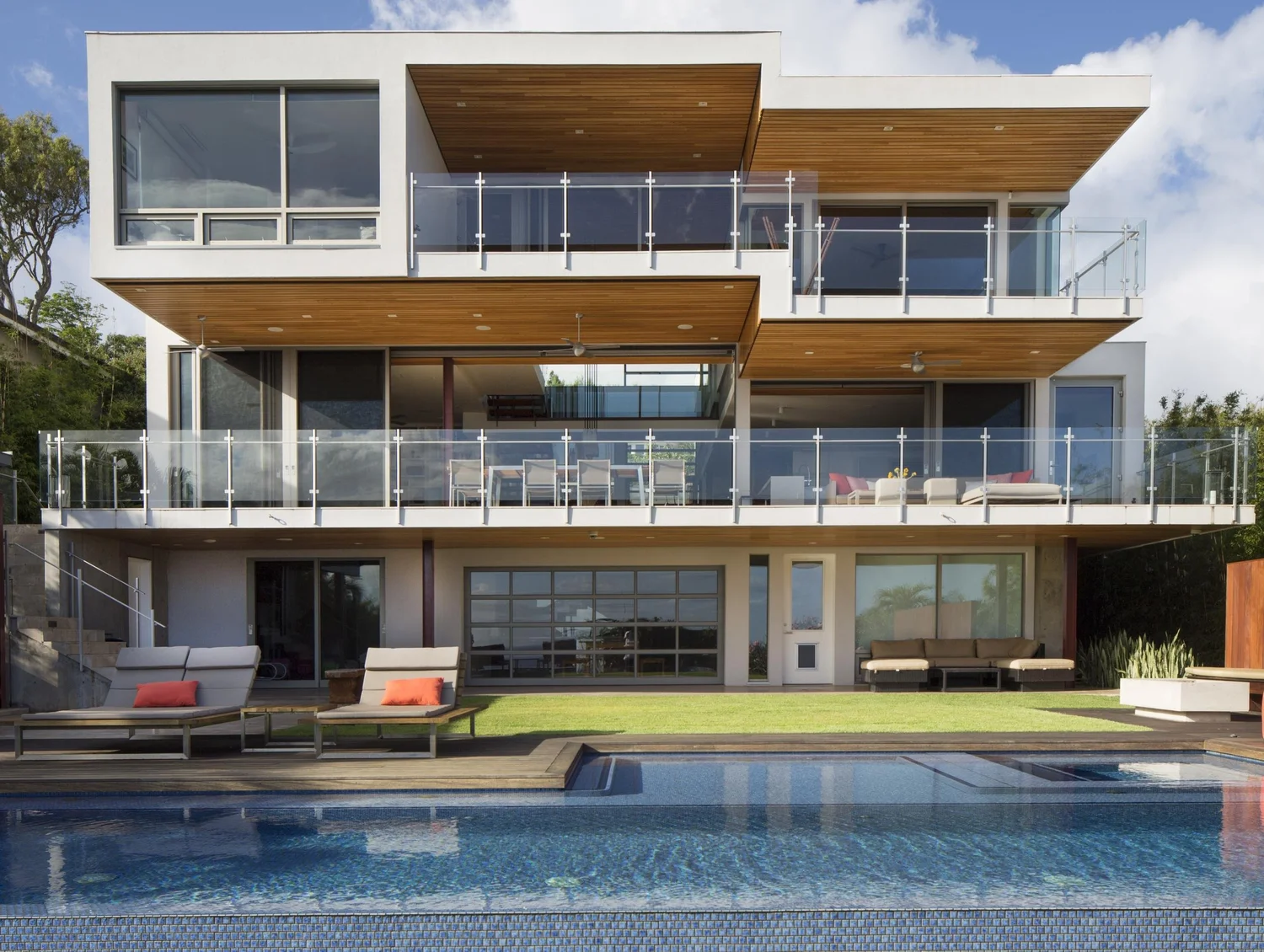 Three-level white house with wood-lined cantilevered overhangs and glass railings, seen from the pool