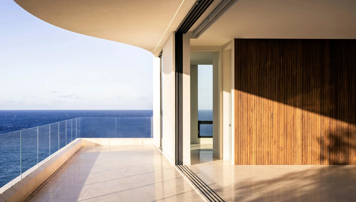Curved lānai with glass railing open to the ocean, low sun across a slatted wood wall