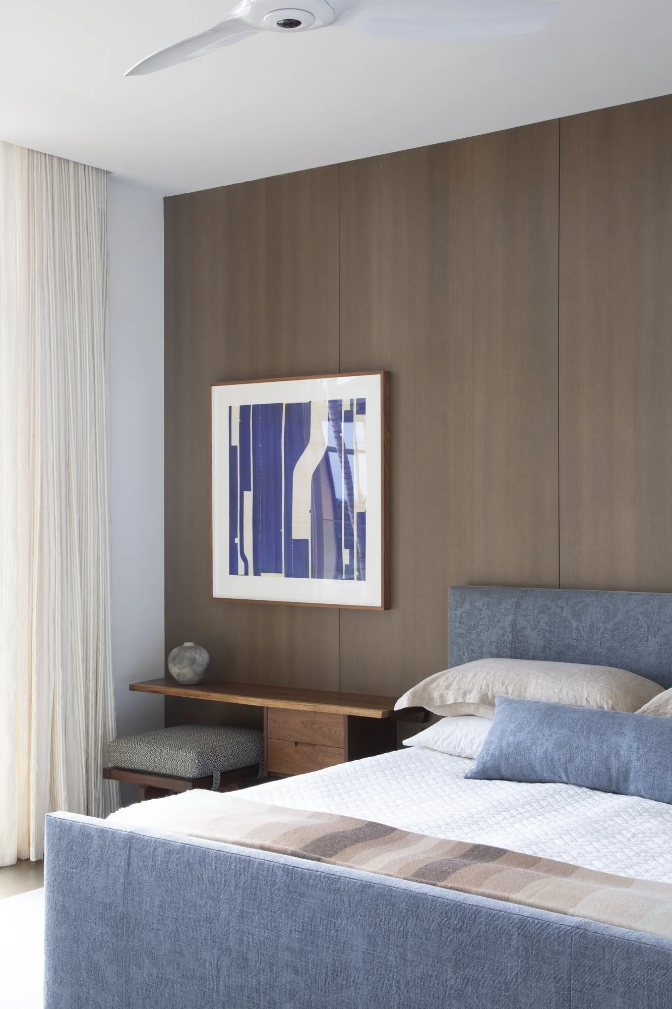 Bedroom with a pale wood-panel wall, framed blue print and sheer curtains