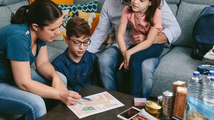 A family sits on a sofa with emergency supplies set on the coffee table while they talk and plan an evacuation route.