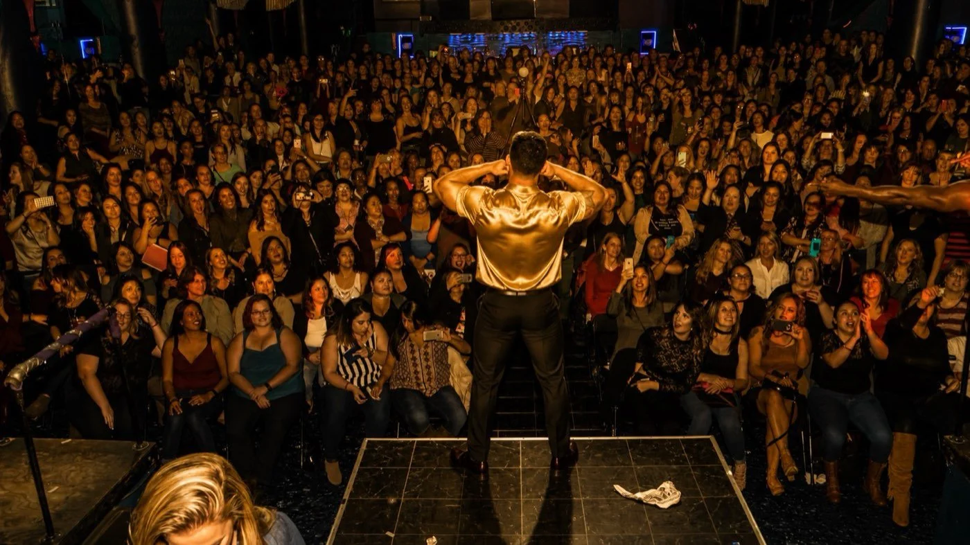 A performer in a gold shirt on stage facing a full room of women