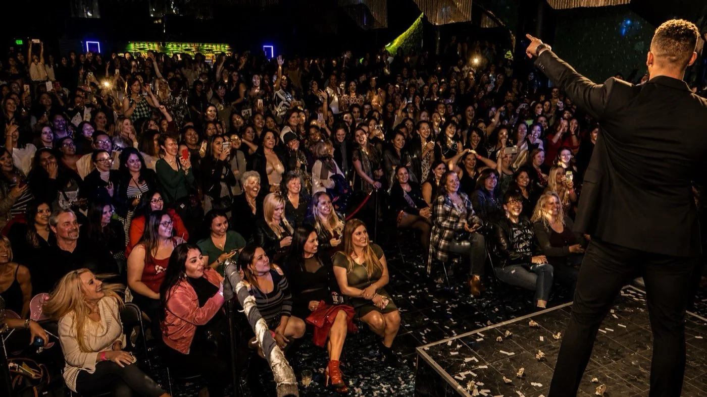A performer in a suit points out over a packed room of women