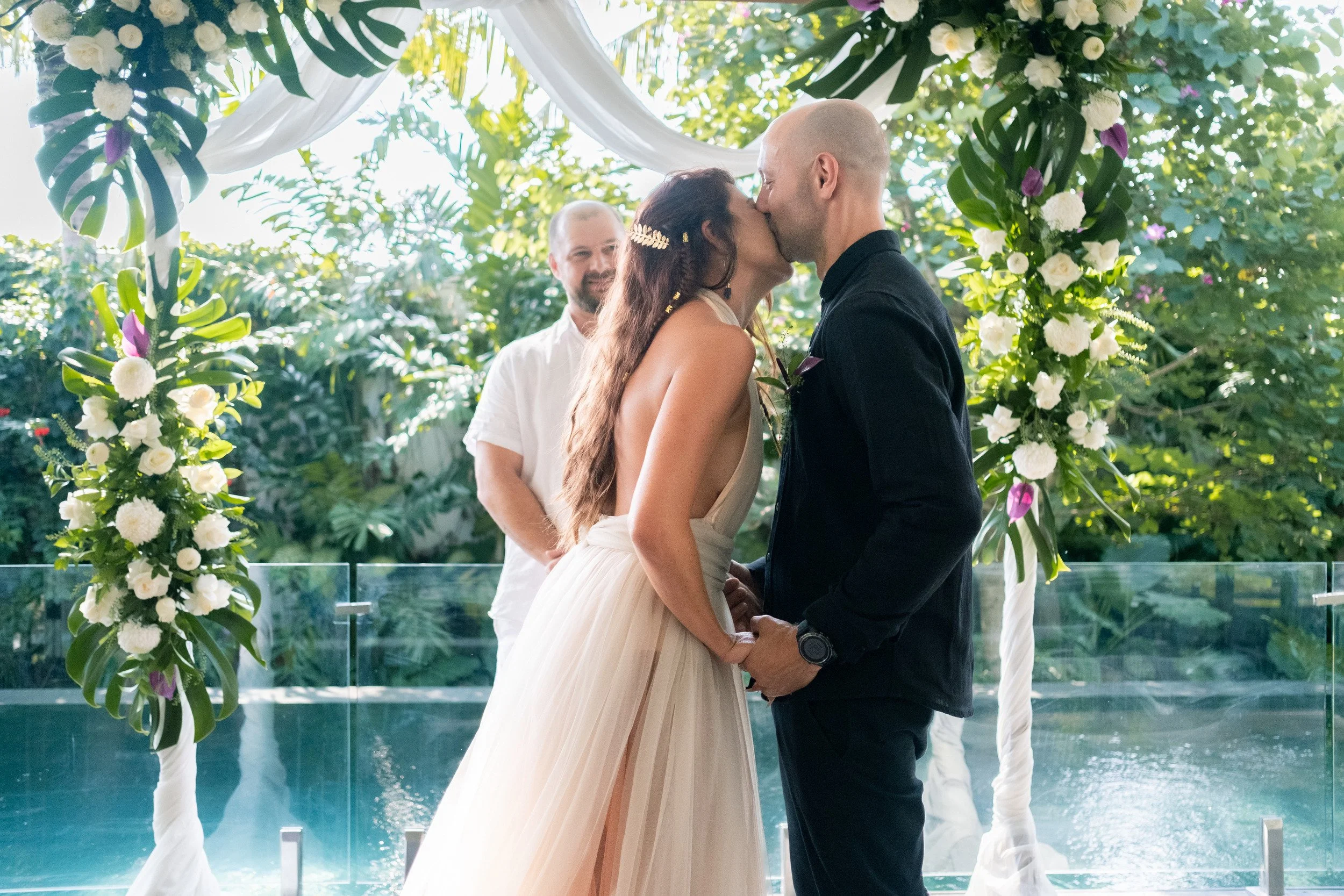 A wedding couple beneath a white floral arch beside Kasianda’s pool