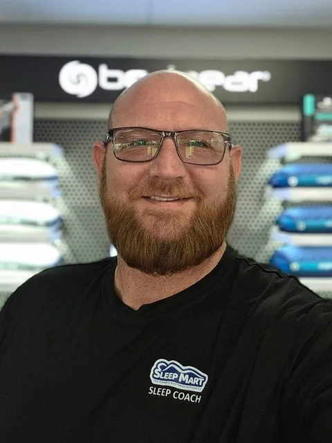 Joe Dixon smiling, wearing glasses and a black Sleep Mart Sleep Coach T-shirt, in front of a Bedgear pillow display wall