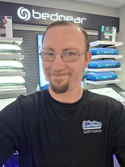 Josh Wyble smiling, wearing glasses and a black Sleep Mart Sleep Coach T-shirt, in front of a Bedgear pillow display wall