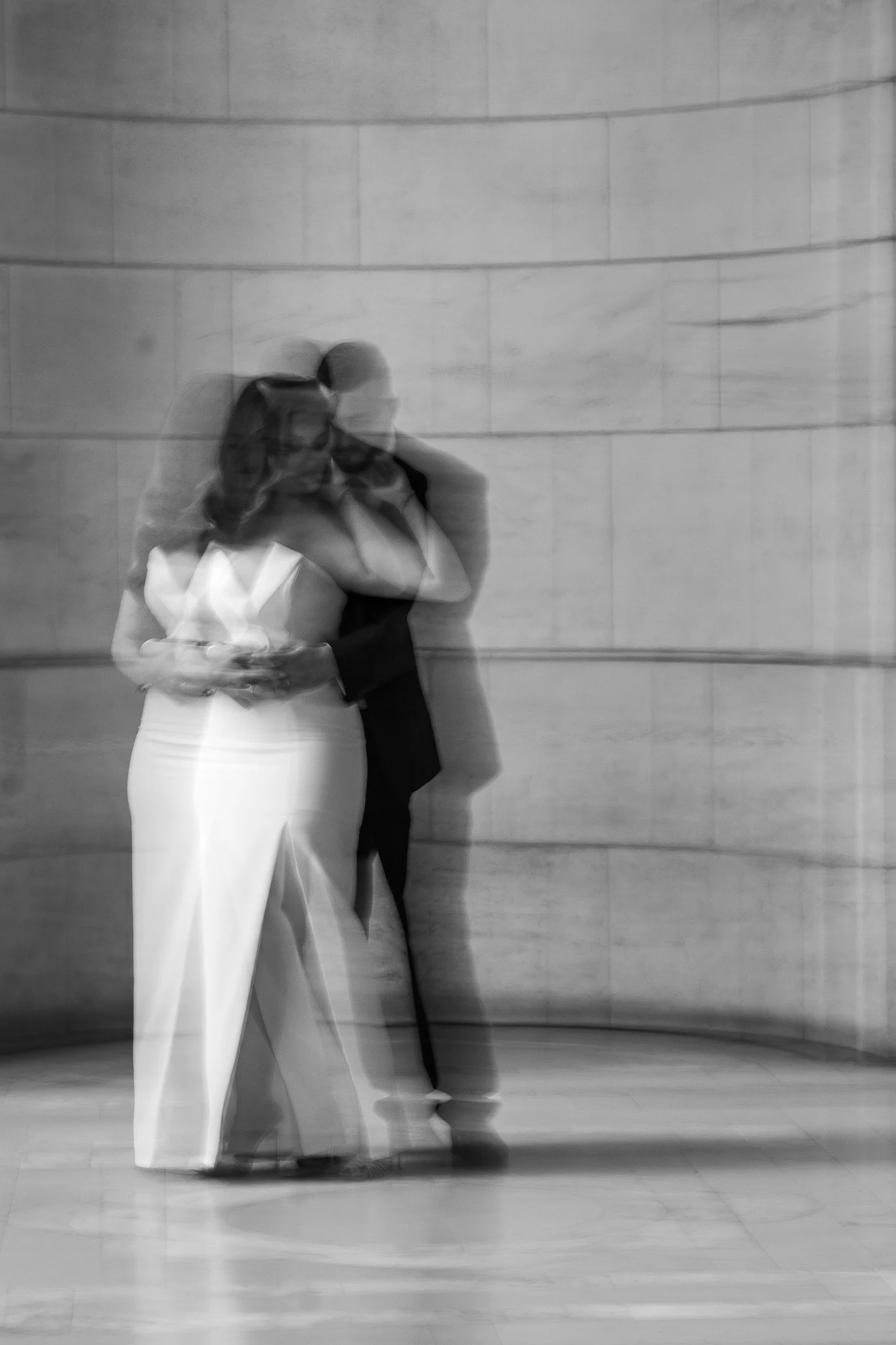Black and white portrait of a couple embracing with intentional motion blur against a curved stone wall.