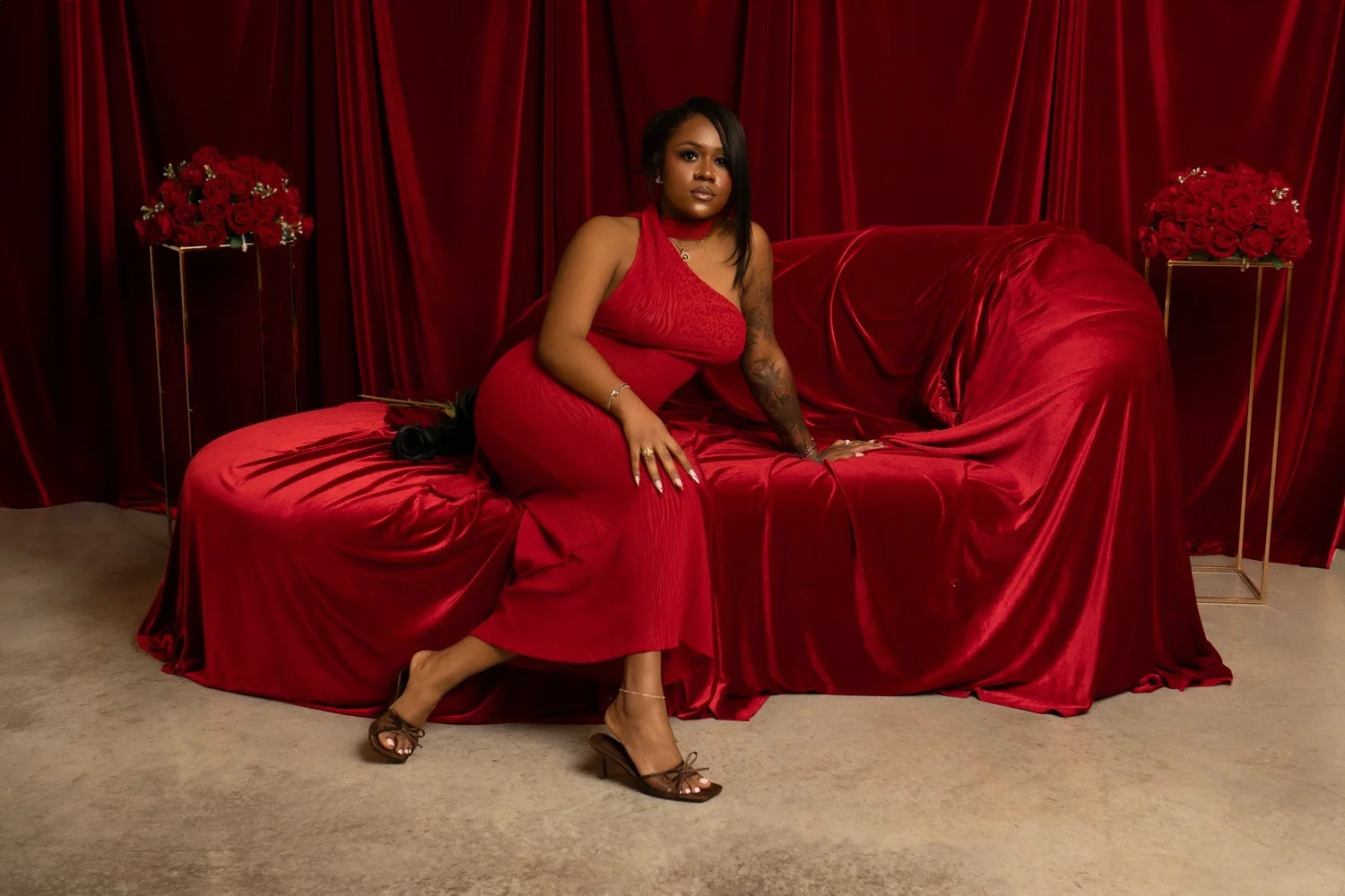 A woman in a red dress seated on a red velvet couch against matching red curtains.
