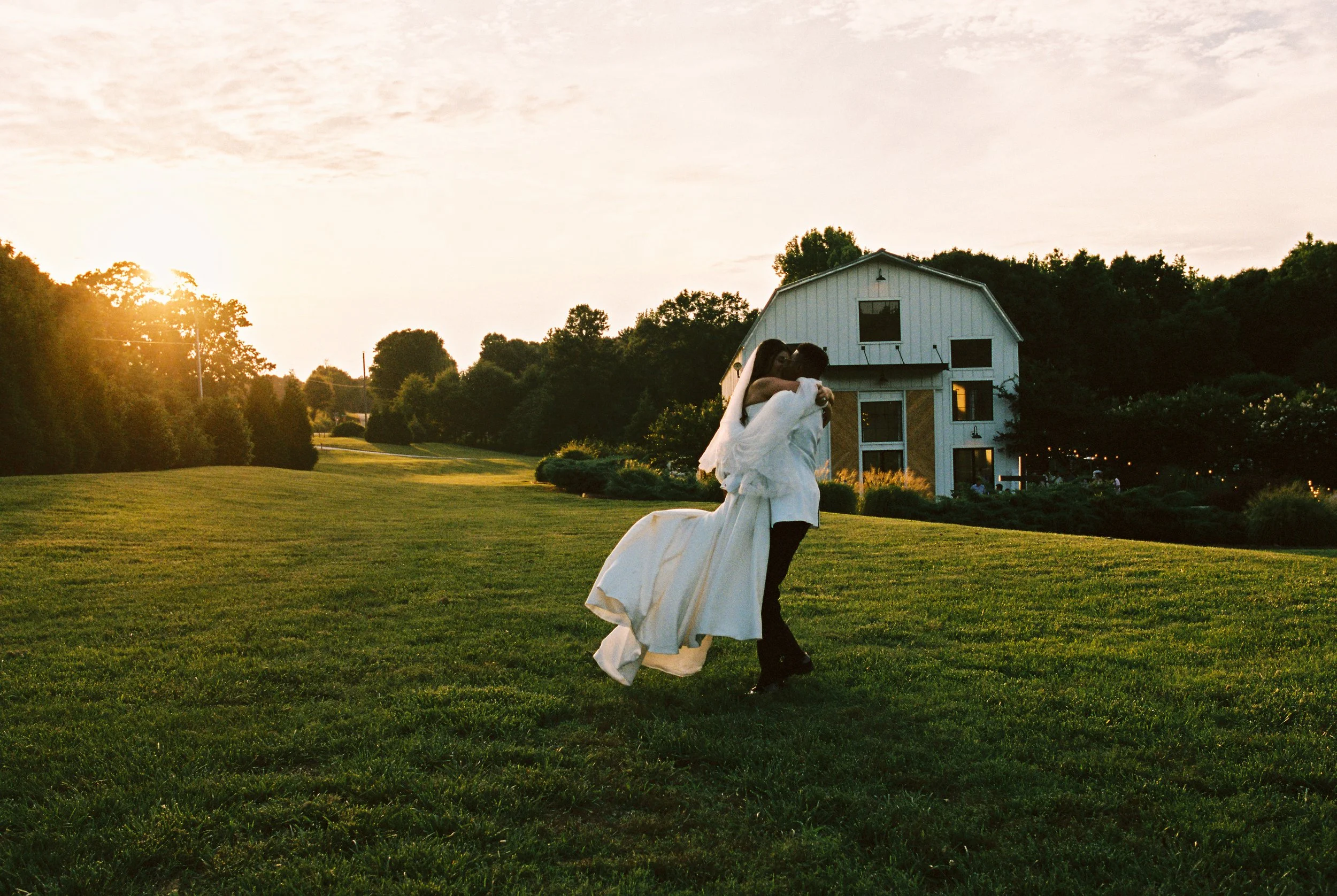 Couple embracing on the lawn at sunset in front of a white barn at Barn South in Talmo, GA