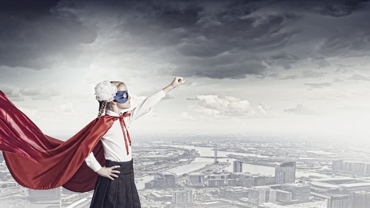 Blonde girl with a red cape and a blue mask and a white flower in her hair, white shirt, and black skirt, standing confidently with one arm outstretched Superman-style in front of a black and white city backdrop.