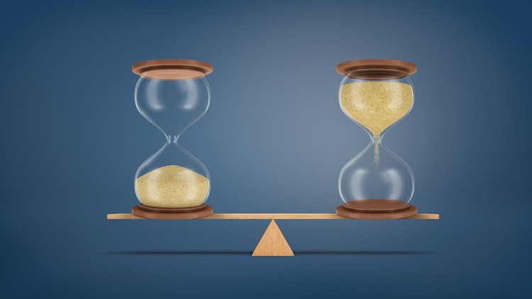 Two hour glasses balancing on a teeter-totter against a navy blue background- one hourglass with sand flowing down from the top and one with sand already filling the bottom.