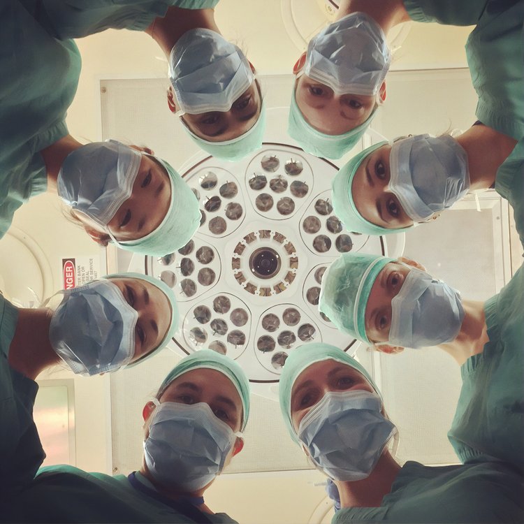 Point of view of a patient looking up and seeing 8 surgeons huddled around, looking down at you in a white room.