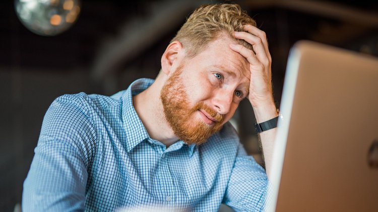 Man with short red hair and a red beard and a blue and white button up dress shirt sits in front of his laptop with his hand on his head in frustration and embarrassment.