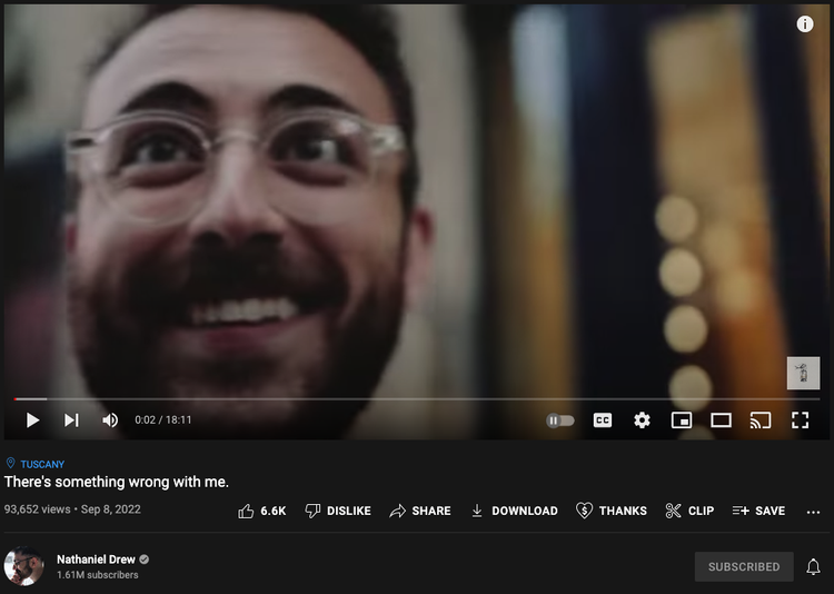Screenshot of Nathaniel Drew YouTube video, "There's something wrong with me". Nathaniel is pictured smiling with brown hair, a beard, and glasses, walking through a city. 