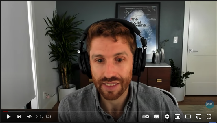 Screenshot of Tristan Harris, with short, brownish red hair and a beard, recording his podcast with headphones on and "The Social Dilemma" movie poster in the background.